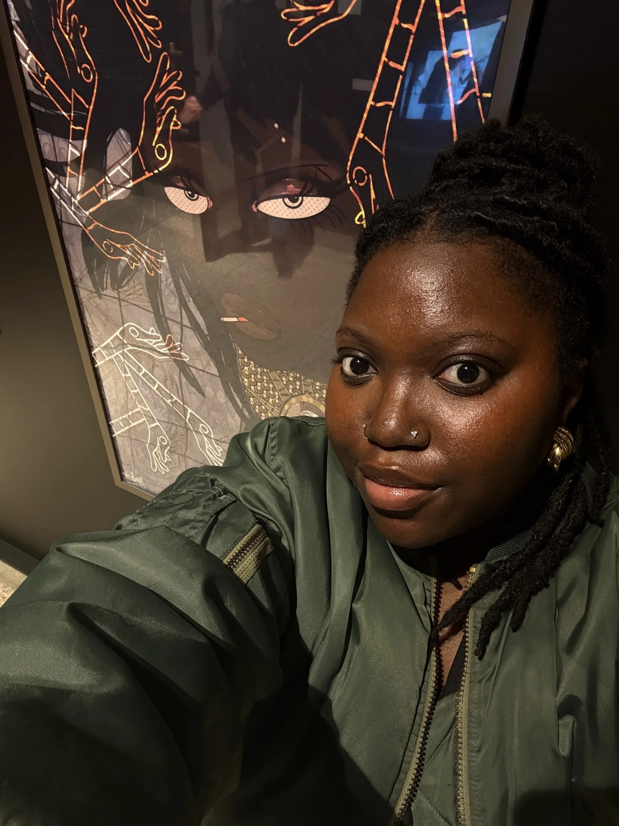 A woman with dark skin and dreadlocks takes a selfie in front of a framed artwork featuring a stylized female face with detailed eyes and intricate accessories.