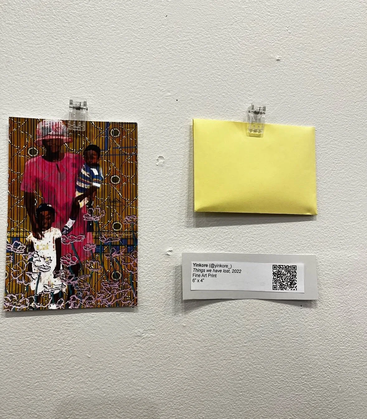 A photograph of two art pieces on a white wall. On the left is a colorful, abstract portrait of a woman with two children, with intricate line patterns. On the right is a small, plain yellow wrapped item pinned to the wall. Below the artwork is a label indicating the artist 'Yinkore,' the title 'Things we have lost,' the year 2022, and the medium 'Fine Art Print,' measuring 6 inches by 4 inches.