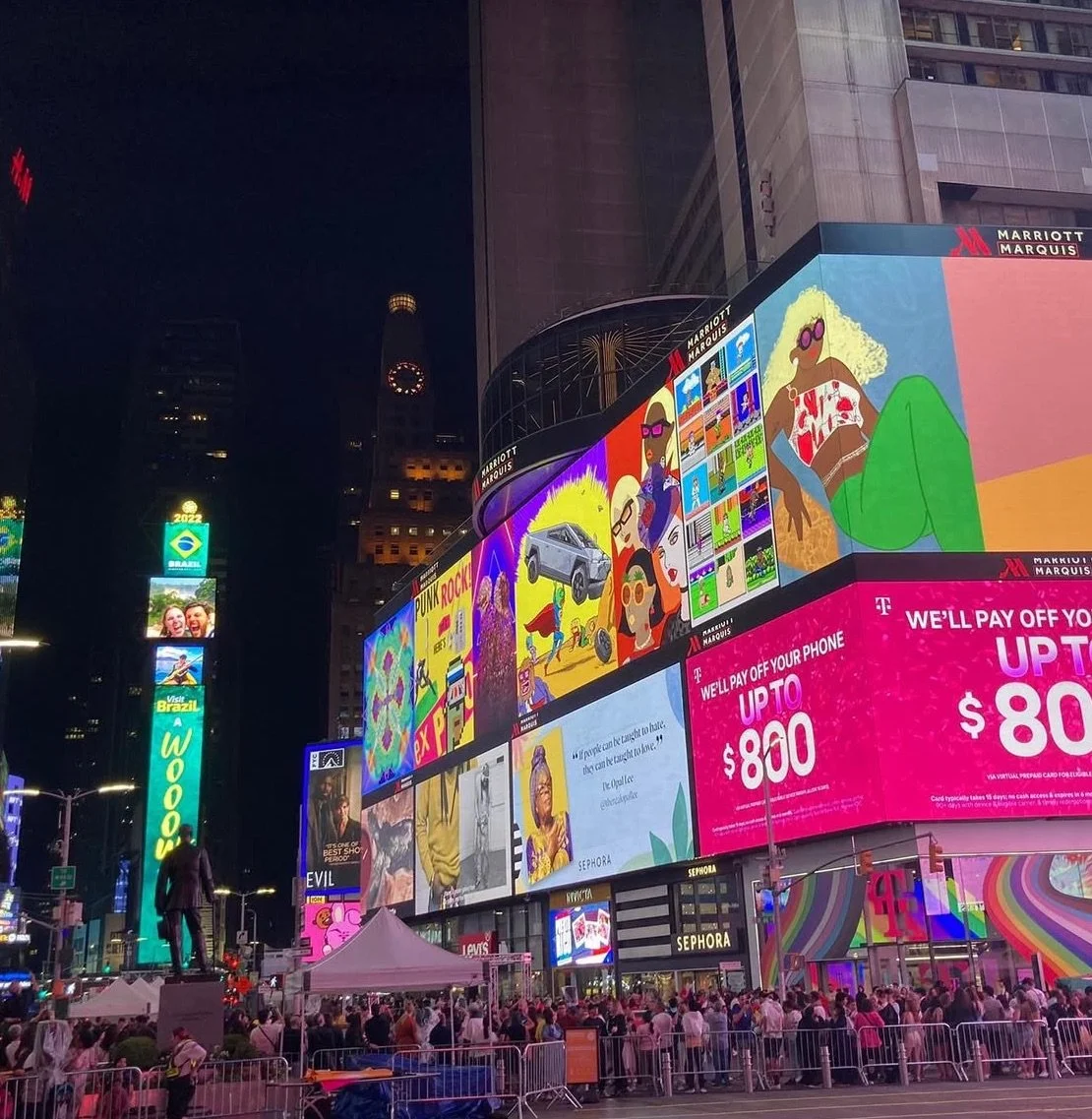 Bright digital billboards in Times Square at night with advertisements, a large crowd, and a statue of a person in the foreground.