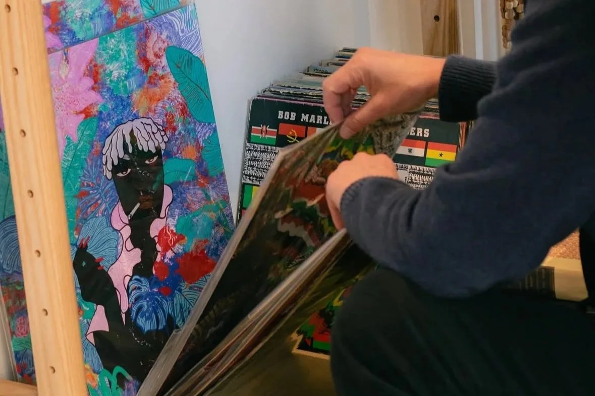 Person flipping through a vinyl record album in a record store, with colorful artwork featuring a woman with dreadlocks and a paint-splattered background.