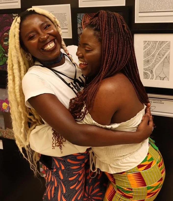 Two women smiling and hugging each other in front of framed artwork make a joyful scene.