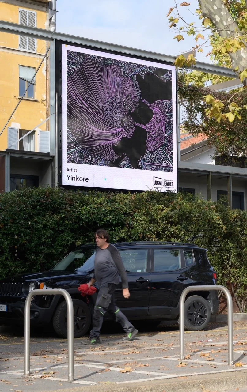 A large digital billboard displaying an abstract, line-art style illustration of a person with flowing hair, purple accents, and intricate details. Below the artwork, text shows the artist's name, Yinkore, and indicates it is part of a local art initiative. A man wearing gray and black clothing is walking past a black SUV parked in front of the billboard on a street with fallen leaves and green shrubs, with yellow and cream-colored buildings and a tree with yellowing leaves in the background.