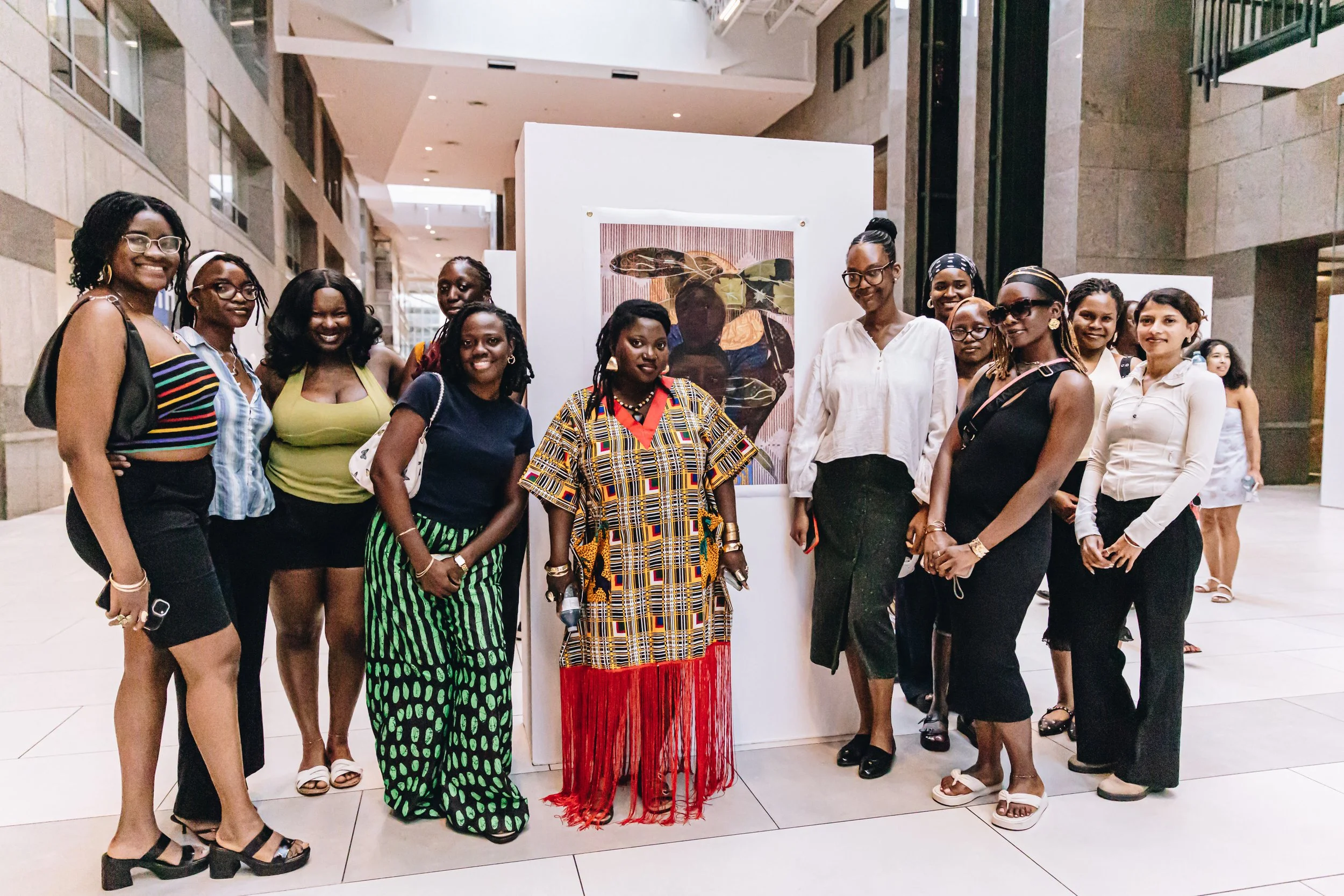 A group of women at an art exhibition, standing in front of artwork displayed on a white panel inside a large, well-lit building.