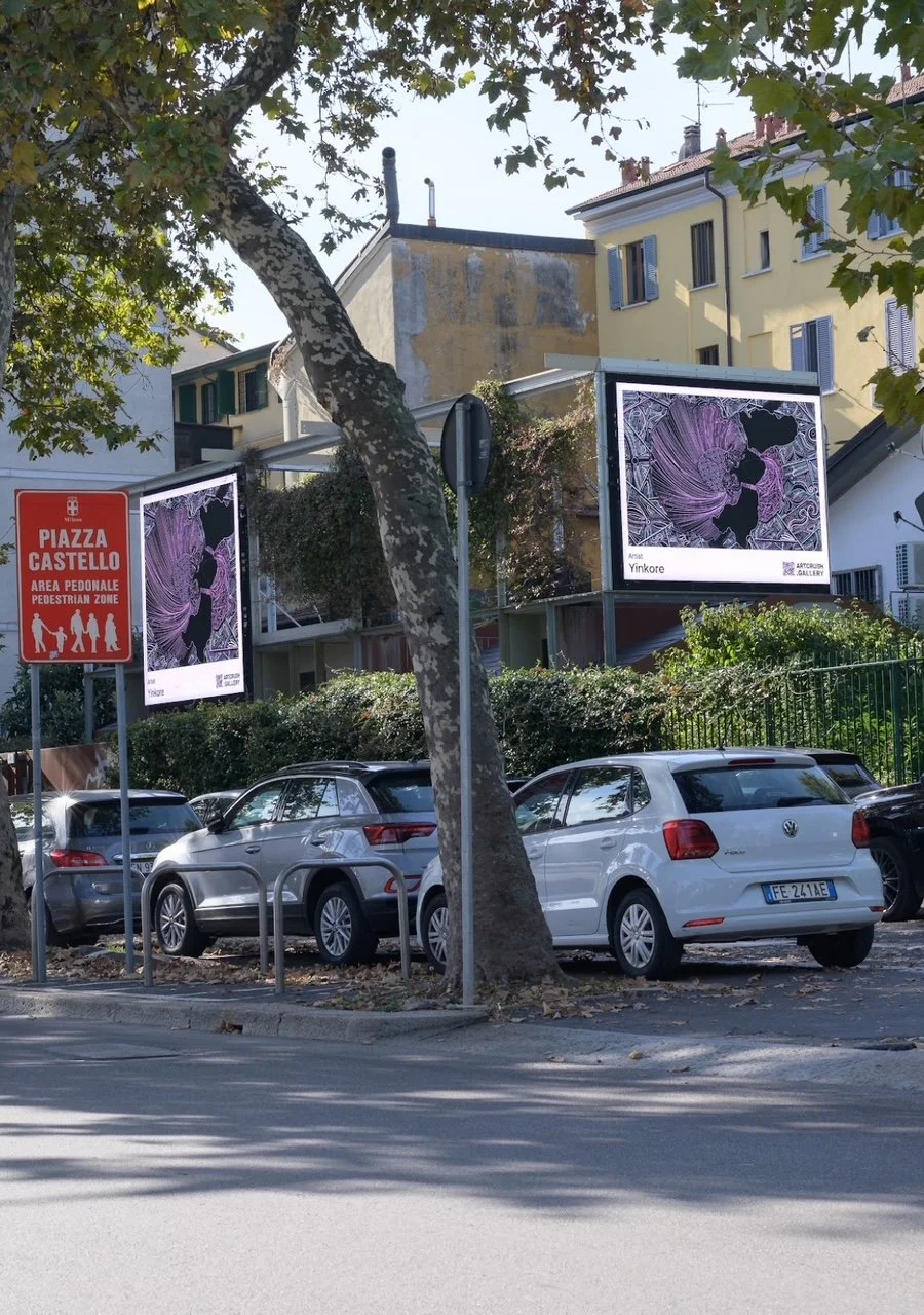 A street scene in Italy with cars parked along the curb, a pedestrian zone sign, and two digital billboards displaying artwork by Yinkore. Trees and buildings are visible in the background.