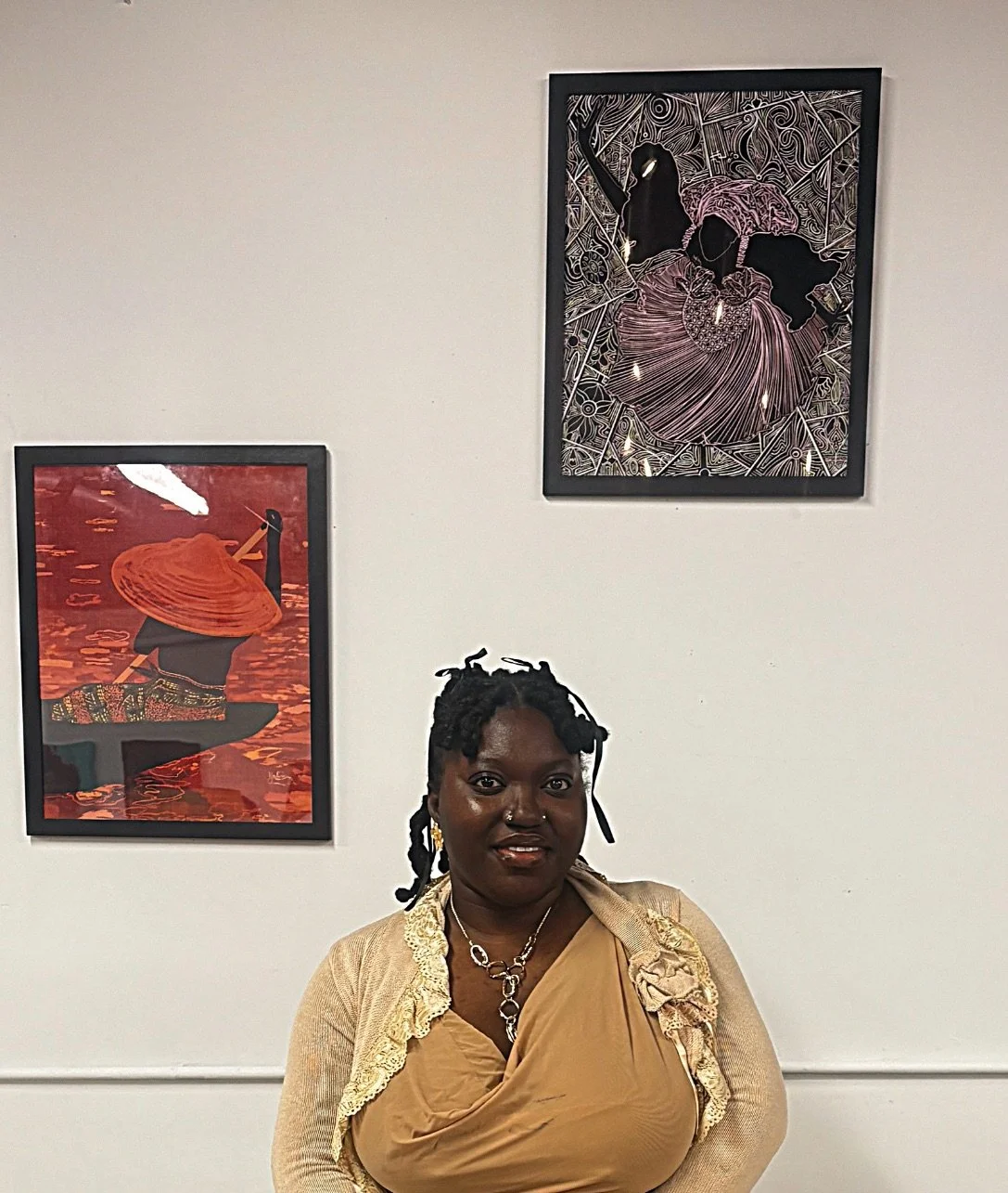 A woman with dark skin and dreadlocks wearing a tan dress and jewelry, standing in front of a white wall with two framed art pieces.