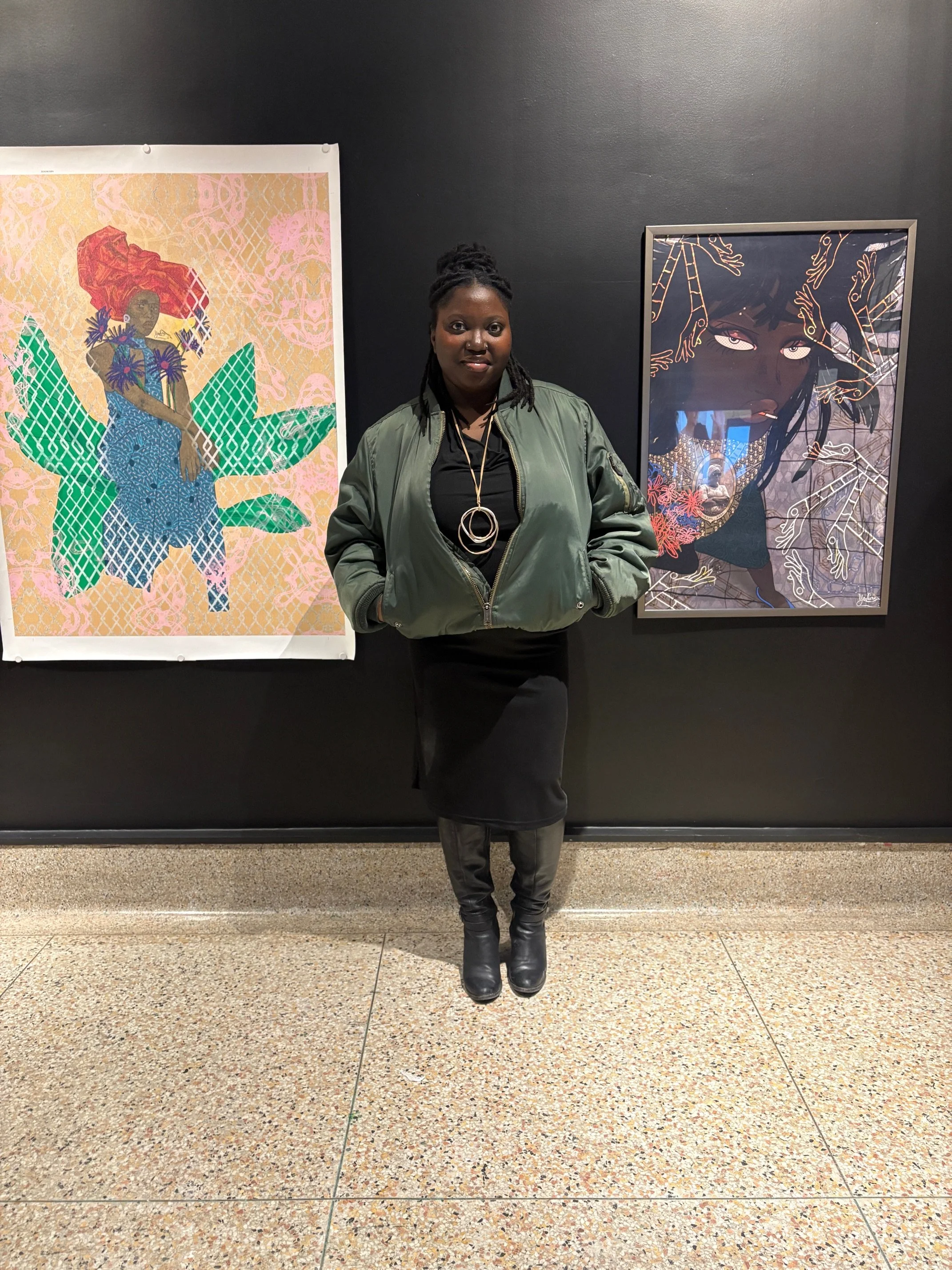 A woman standing in front of two colorful artworks in an art gallery. She is wearing a green bomber jacket, black dress, and black boots, and is smiling at the camera.
