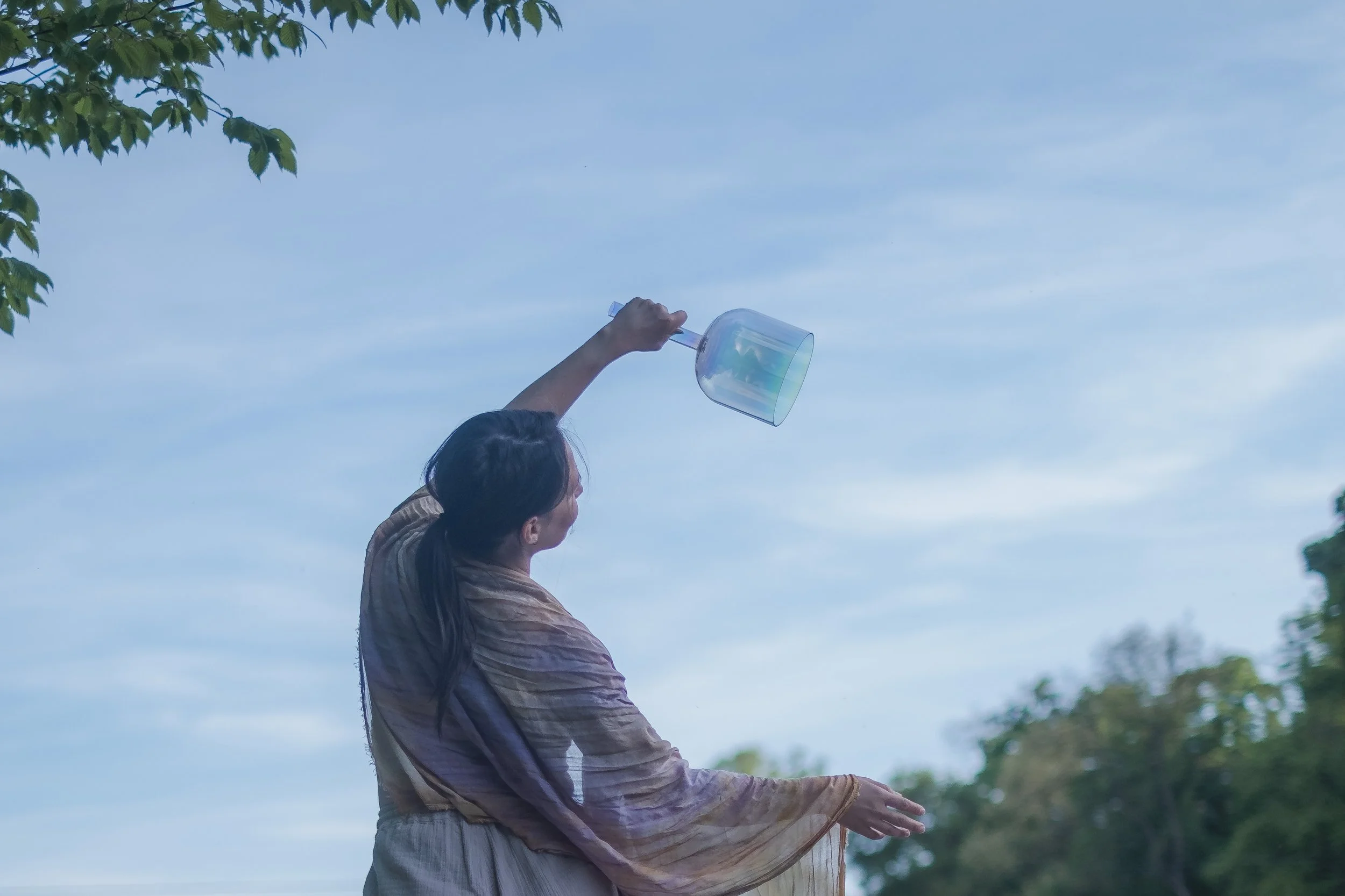 A person with long dark hair stands outdoors under a blue sky, holding a transparent iridescent crystal singing bowl with a stick handle, reaching upward towards the sky.