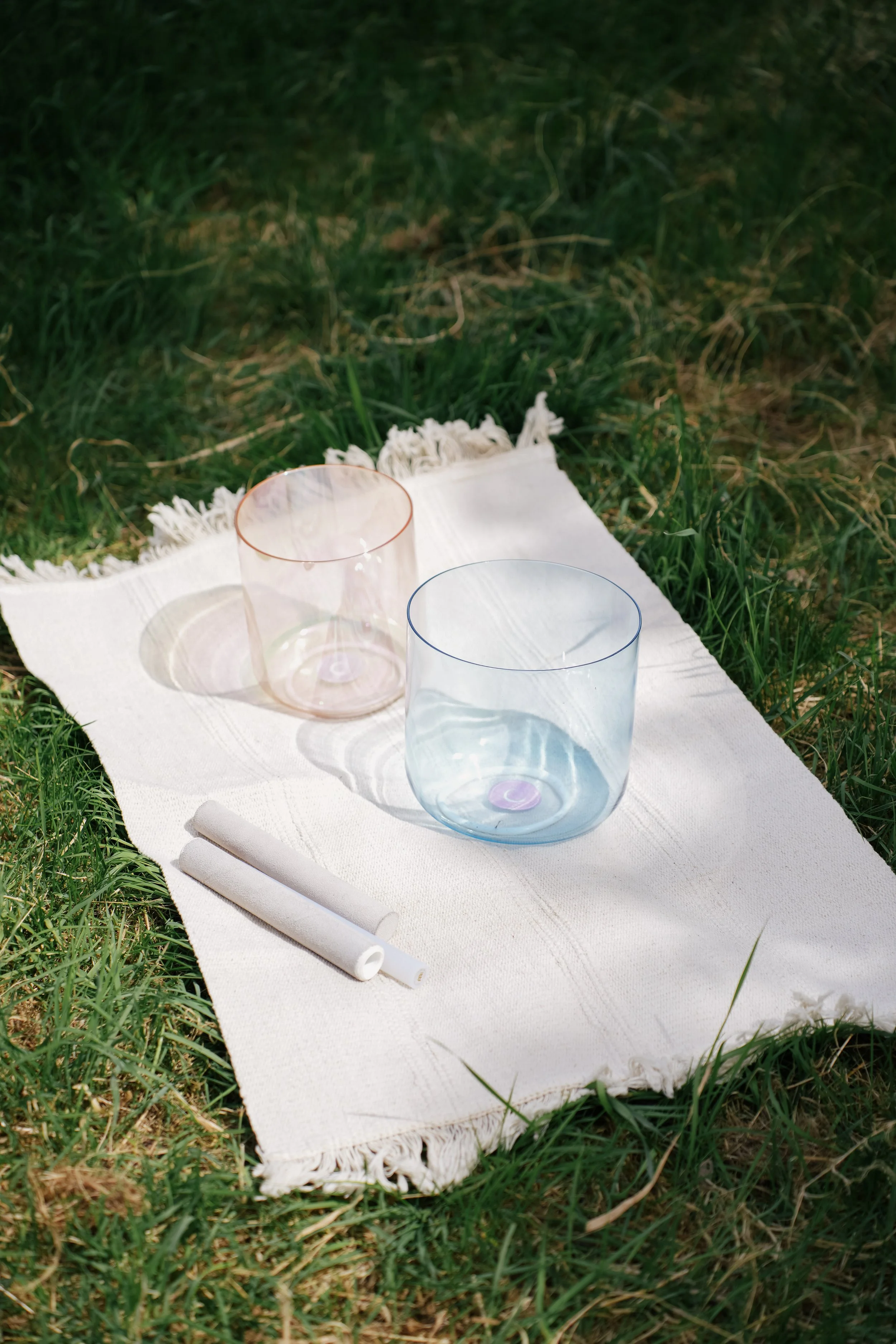 A white textured blanket with fringes spread on green grass outdoors, with two translucent crystal singing bowls, one pink and one light blue, placed on it, along with a white rolled-up paper or napkin.