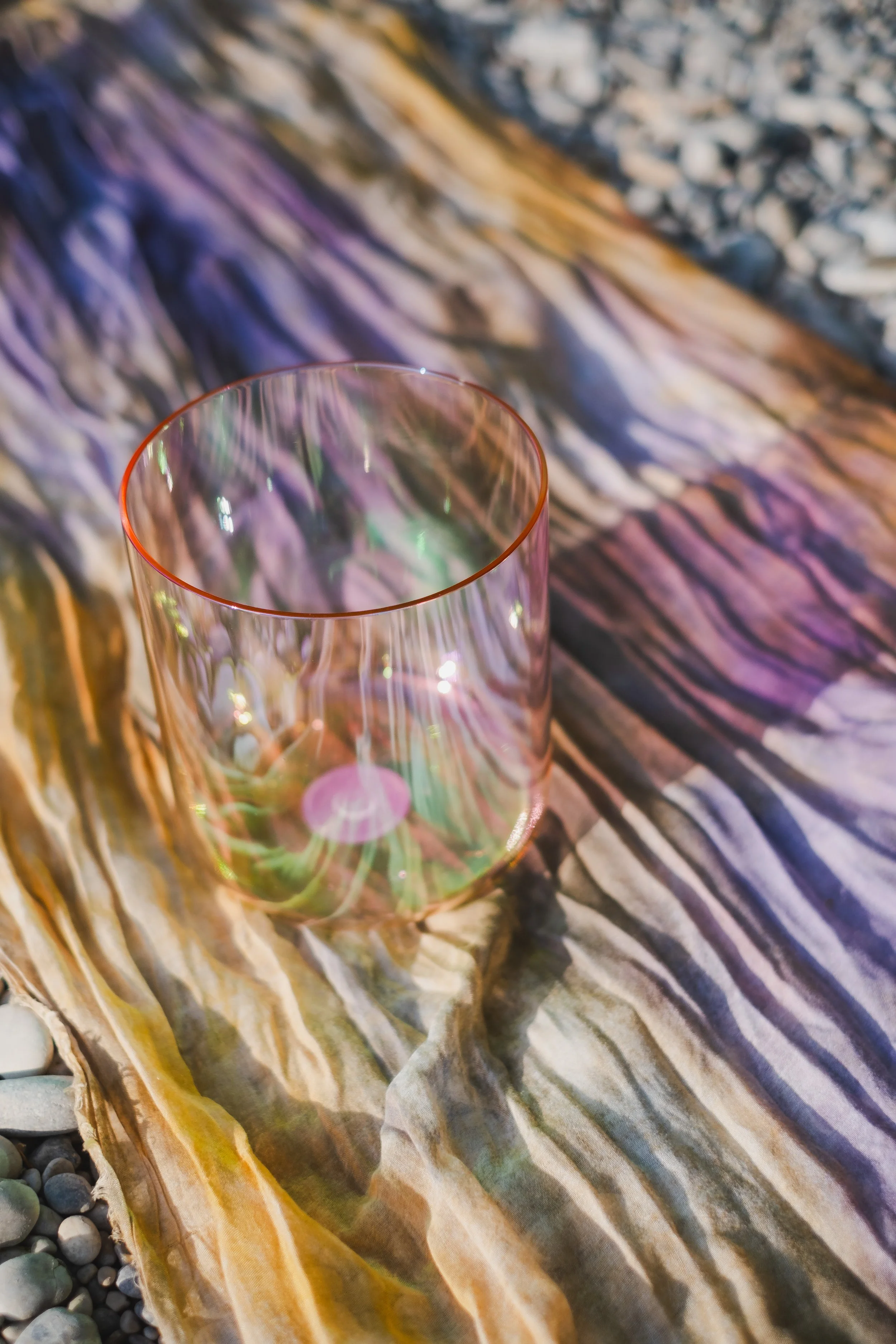 A pink translucent crystal singing bowl with a rainbow-like reflective surface placed on a rocky and colorful wooden surface.