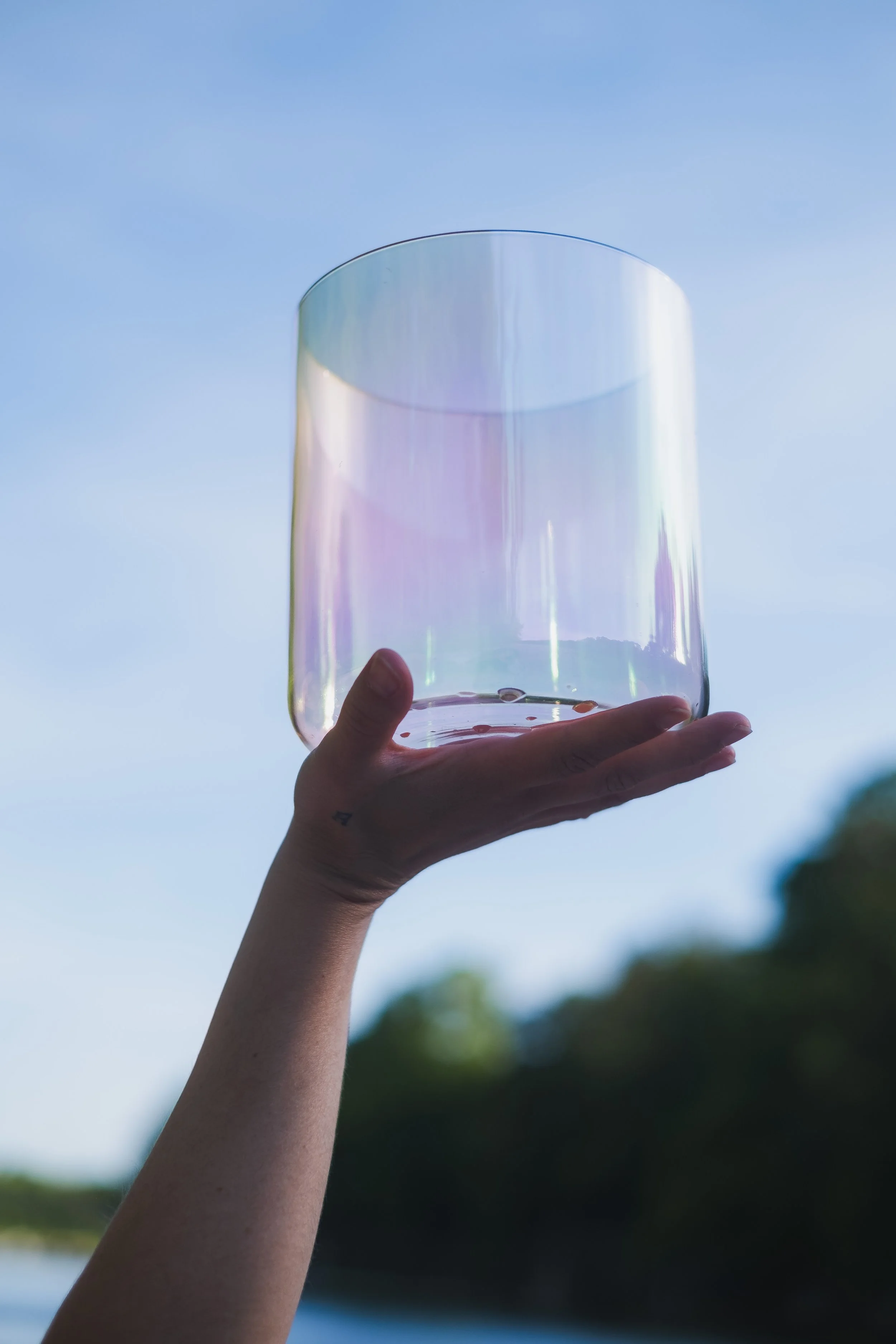 A person holding a clear, iridescent crystal singing bowl up to the sky with a blue sky and blurred trees in the background.