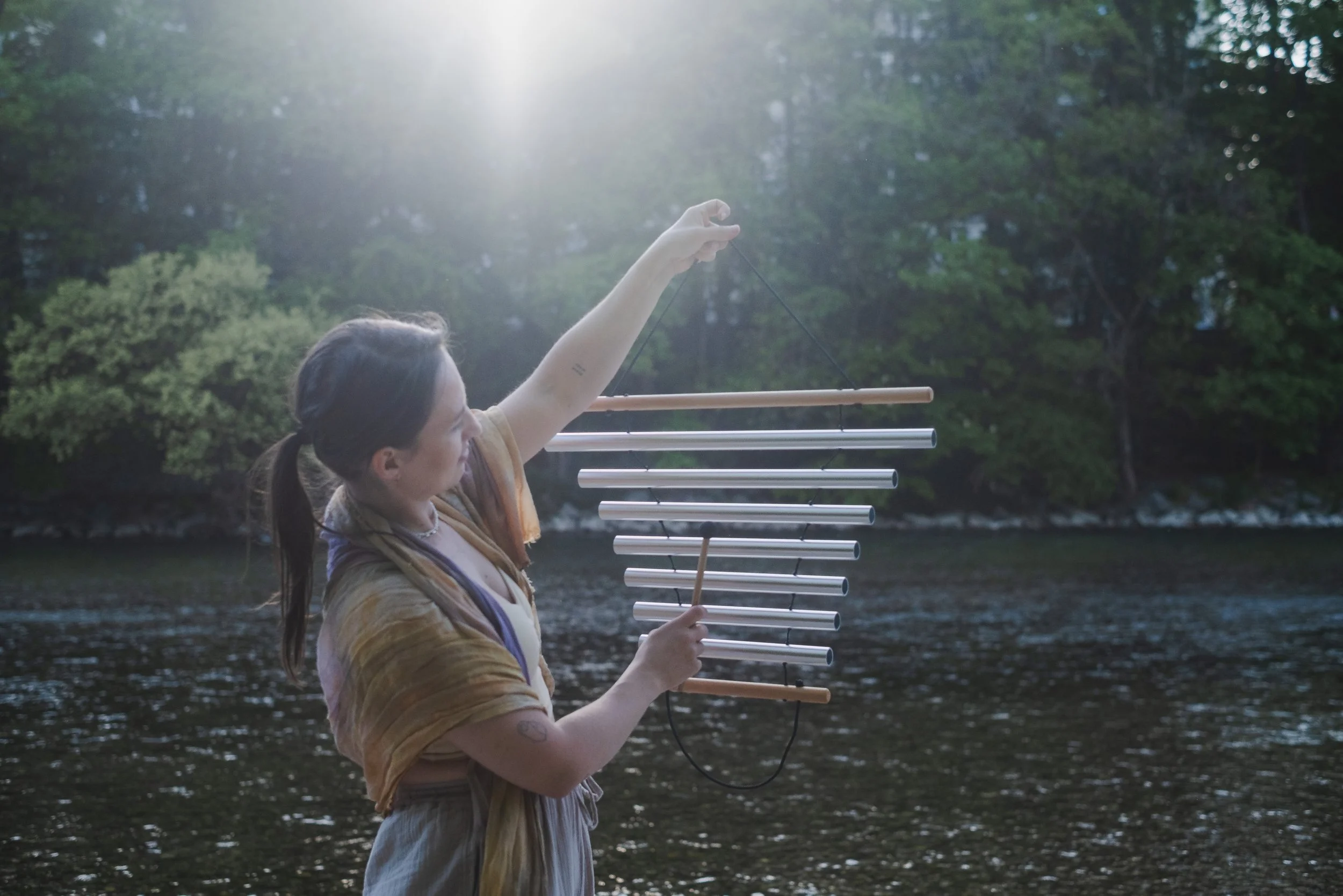 A woman by a river playing a wind chime instrument with metal tubes, holding a stick, outdoors with trees and sunlight