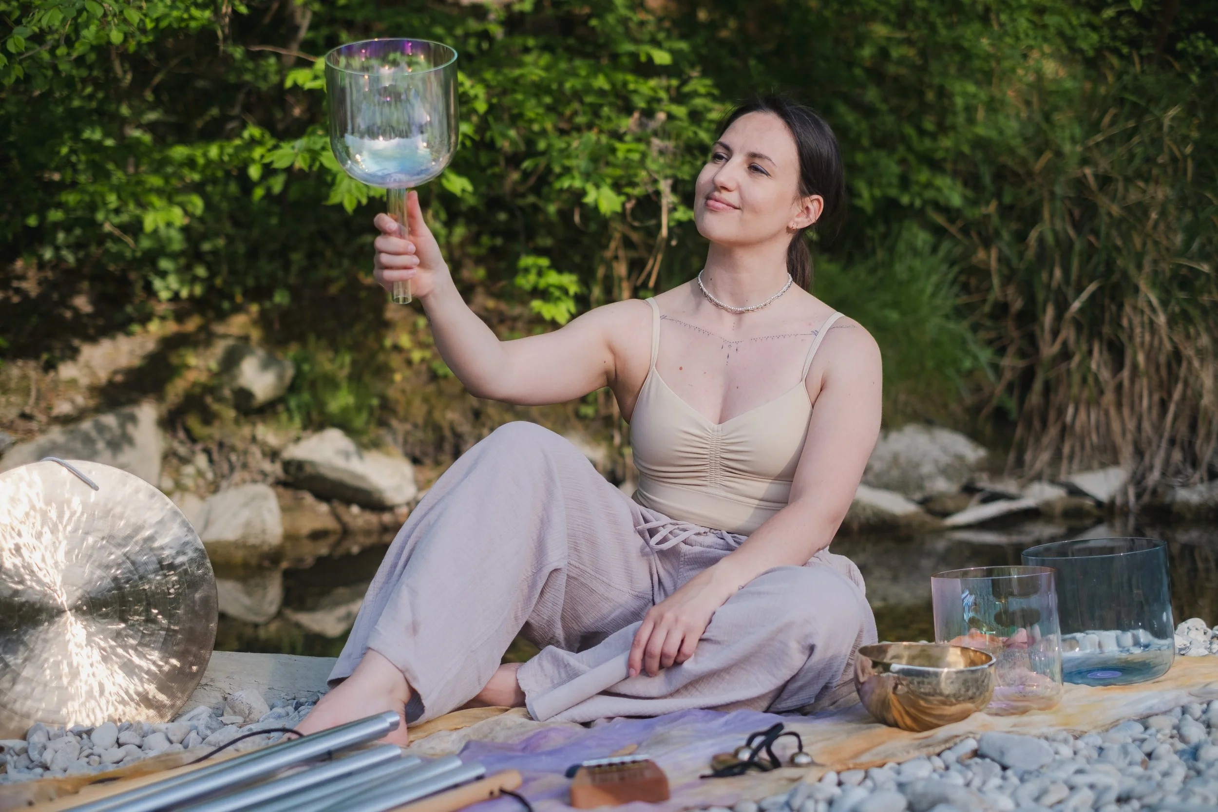 A woman sitting outdoors by a brook, holding a crystal singing bowl and smiling softly. She is dressed in a light-colored top and light pants, surrounded by sound instruments on a blanket, with greenery and rocks in the background.