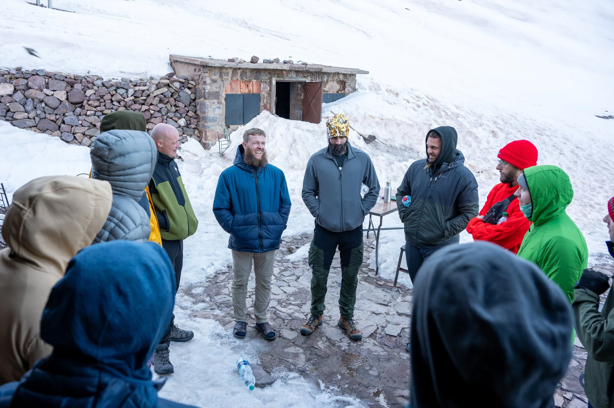 Group of people in winter clothing gathered outdoors on snow-covered ground, standing in a circle and talking, with a small stone structure in the background.