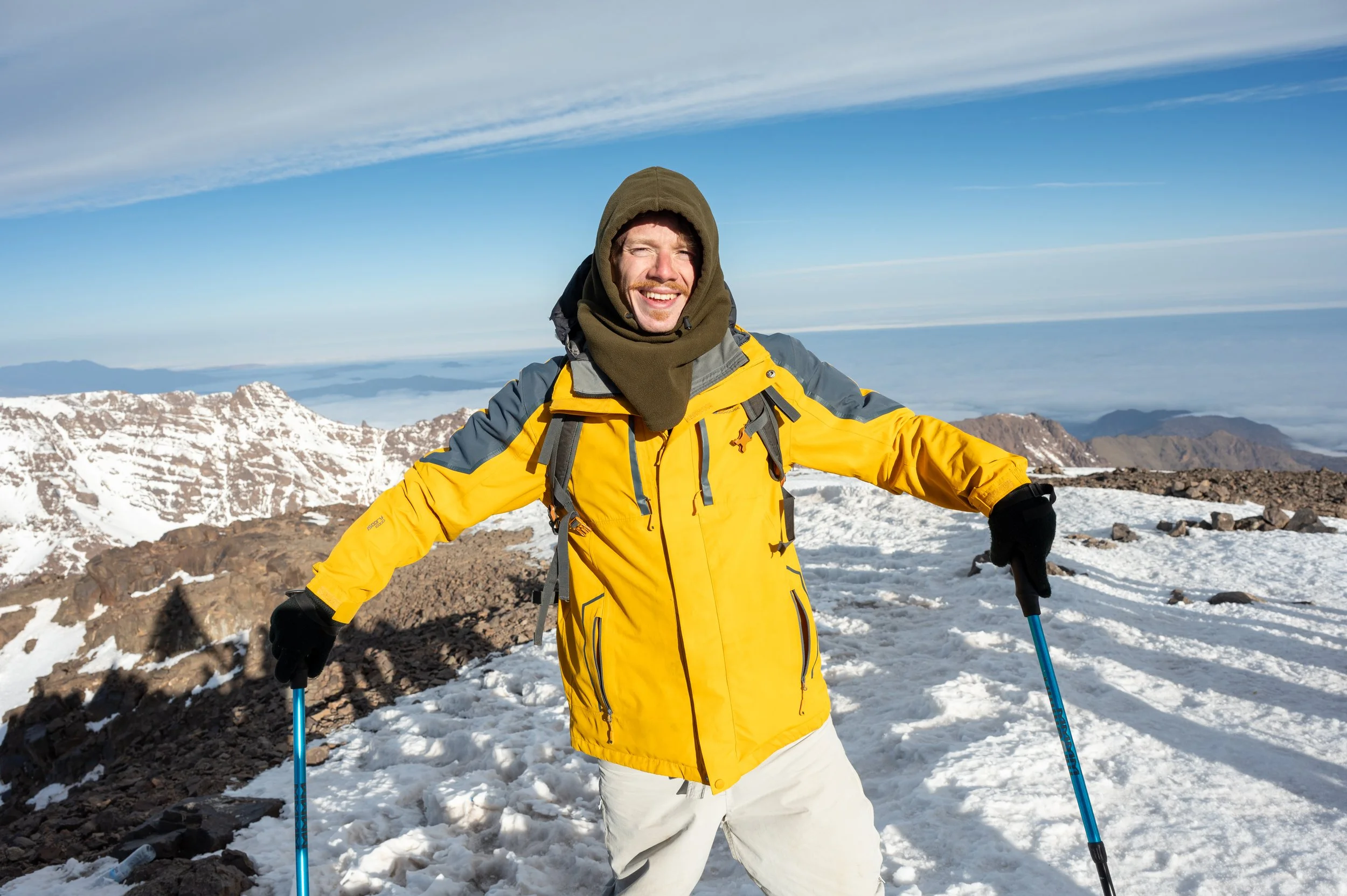 Man wearing yellow jacket and black gloves on snowy mountain with snow-covered peaks and blue sky in the background.