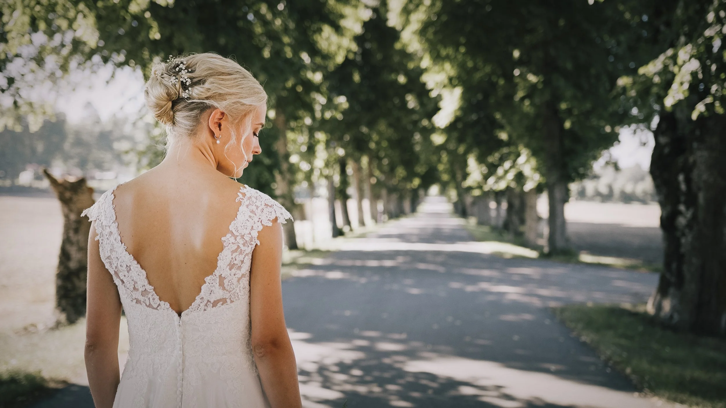 En brud i vit brudklänning med blommor i håret står på en trädkantad allé.