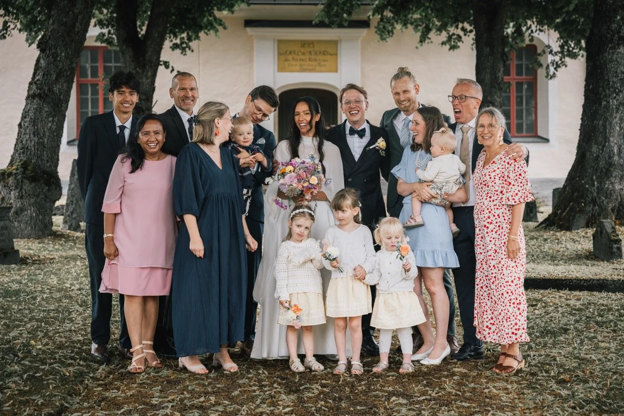 En grupp människor poserar för ett bröllopsfoto utanför en vit byggnad med träd i bakgrunden. Det är en bröllopsbild med brud, brudgummen och familjemedlemmar och barn.