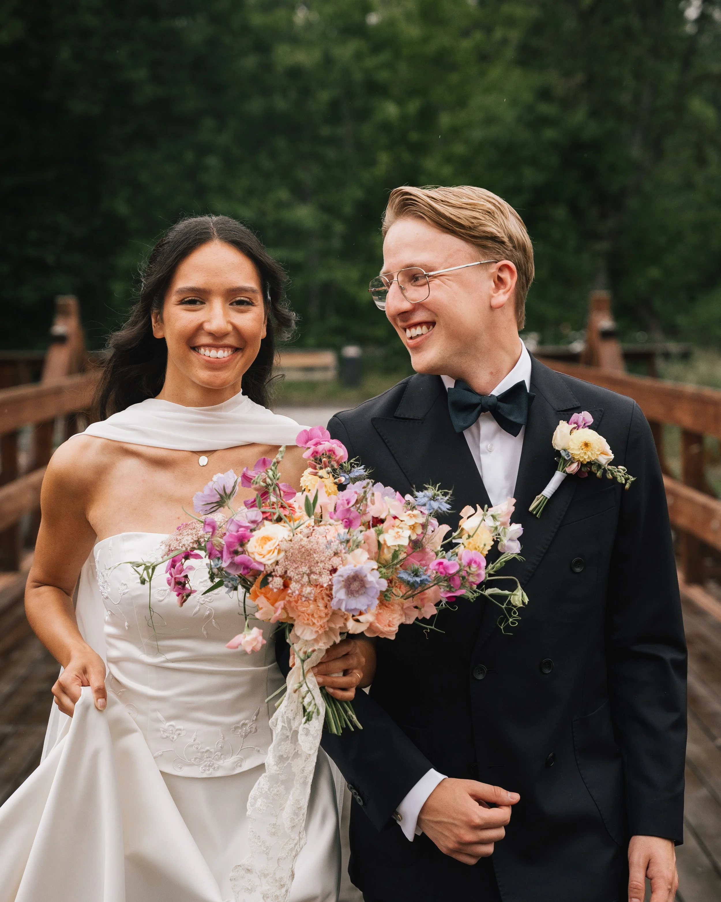 Ett brudpar i formella kläder, kvinnor i brudklänning och man i svart tuxedo, på en träbro i en grön park, håller ett stort blomsterarrangemang.