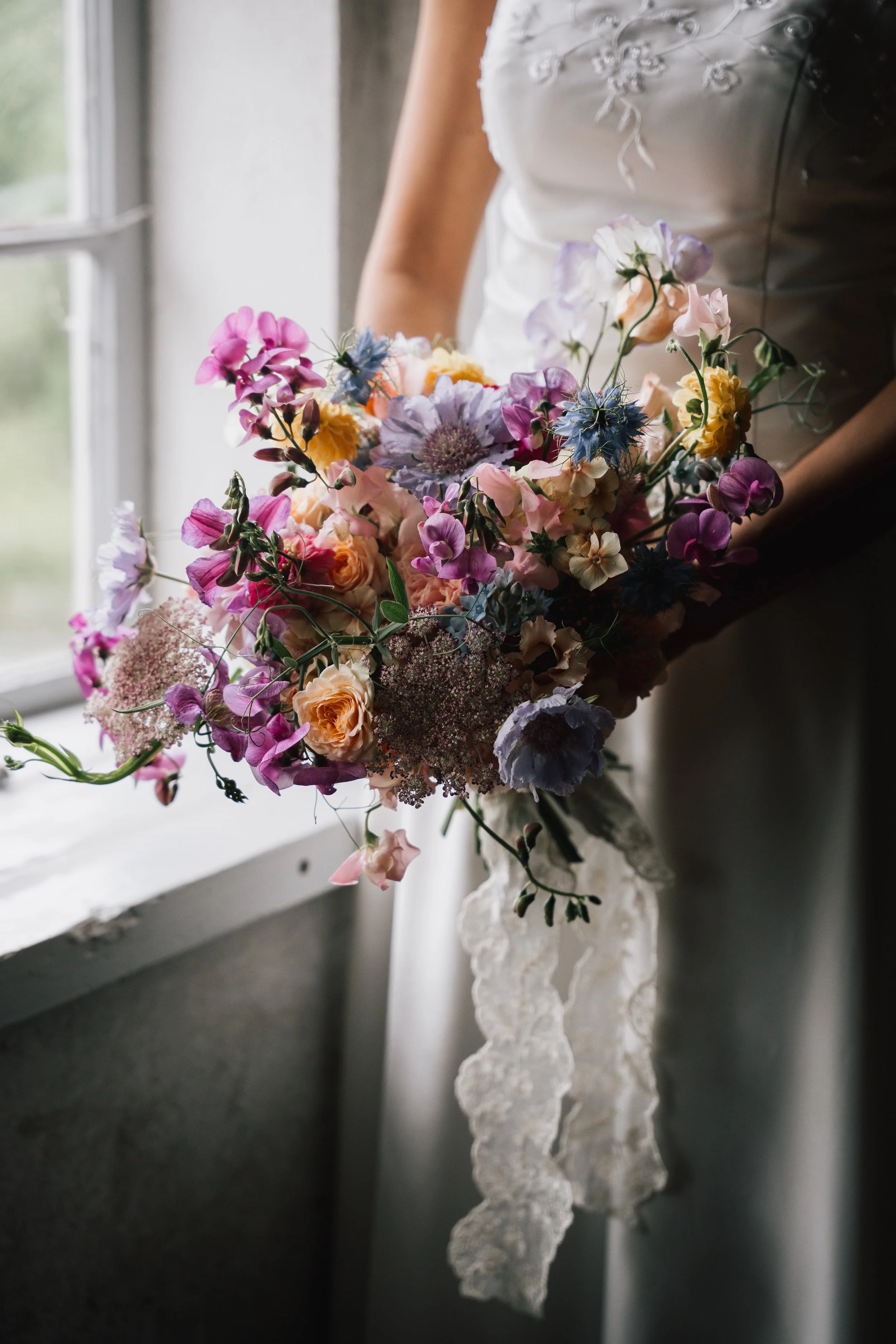 Färgglad brudbukett med olika blommor i pastellfärger, hålls av en person i en bröllopsklänning framför ett fönster.