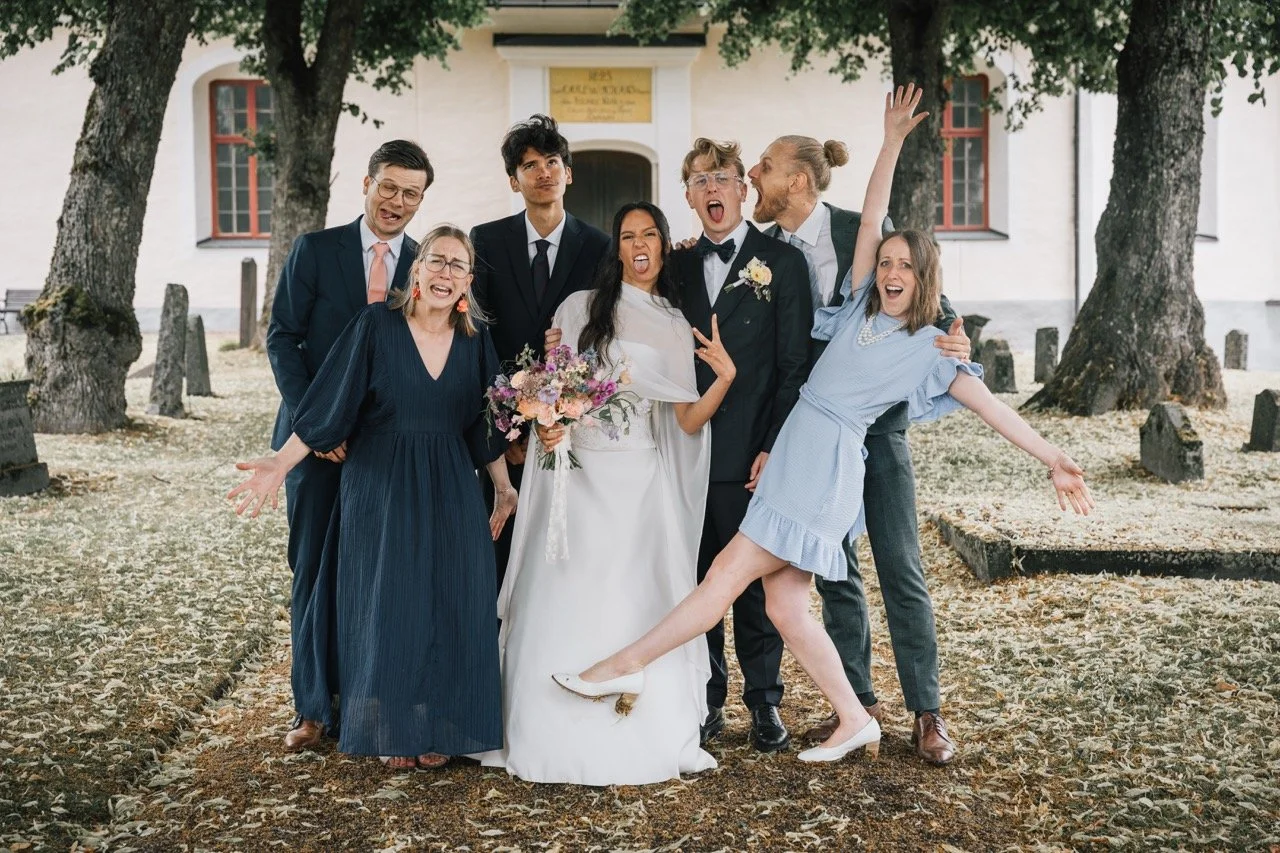 En grupp personer står framför en gammal kyrka, de är klädda för ett bröllop, leende och festliga.
