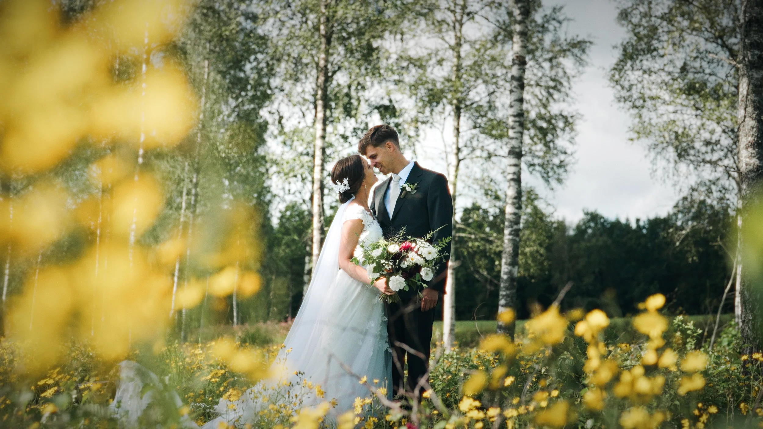 En nygift brud och brudgum står nära varandra i en skog, omgivna av gröna träd och gul blommor.