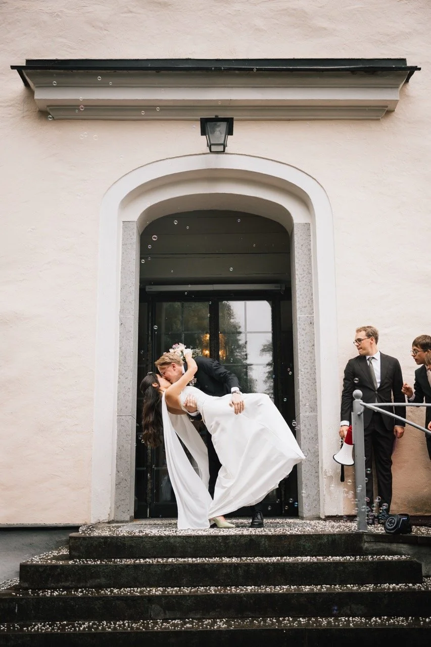 Nygift par kysser framför en ingång till en byggnad, omgiven av gäster, varav några håller en megafon, utomhus.
