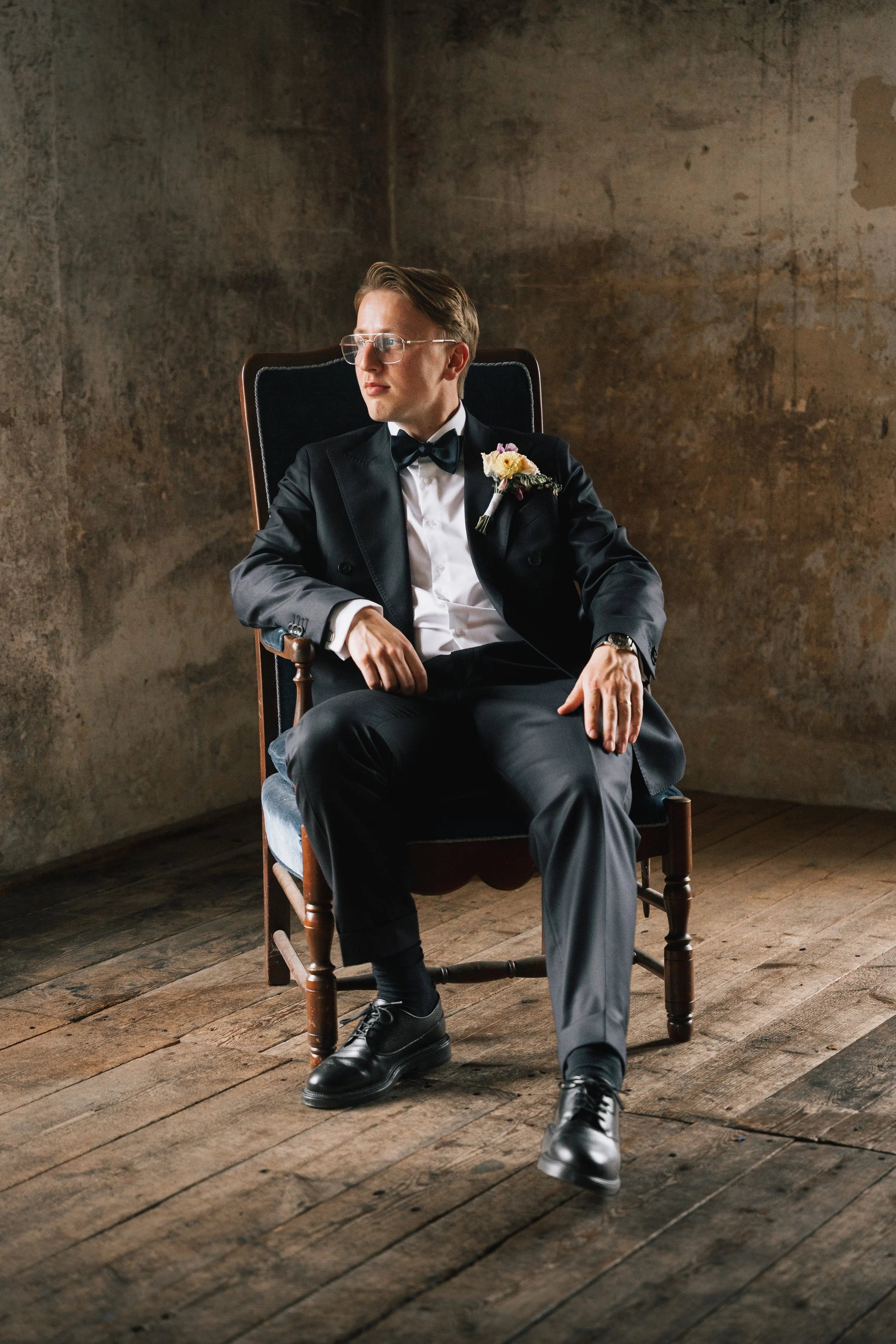 En man i svart smoking med en blomsterboutonnière sitter i en trästol, tittar åt sidan, i ett rustikt rum med trägolv och en grå vägg.