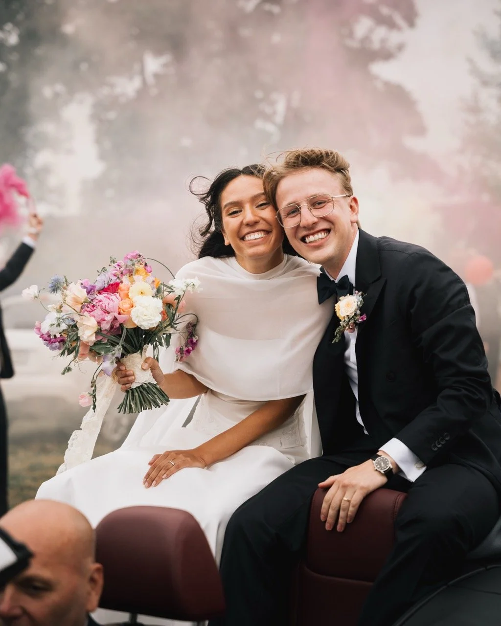 En glad nygift couple på bröllopsdag, kvinna i vit brudklänning och man i svart frack, sitter tillsammans på en häst och vänner som kastar blomblad syns i bakgrunden.