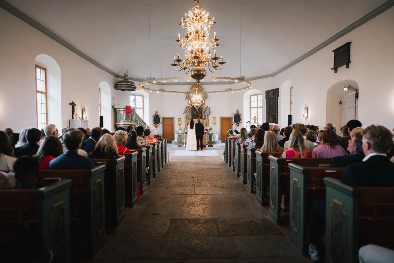 En brud och en brudgum står framför ett altare under en vigselceremoni i en kyrka, omgiven av gäster som tittar på.