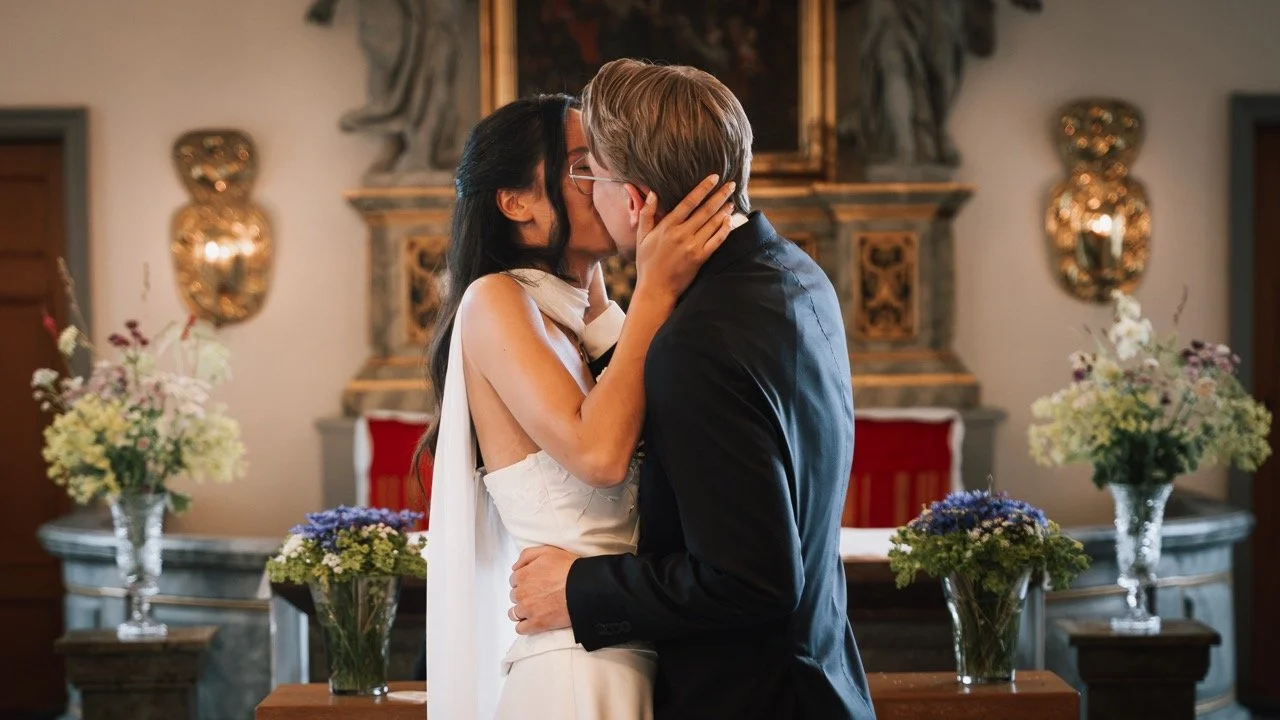 Ett par kysser varandra i en ceremoniell miljö med blommor och konstverk i bakgrunden.