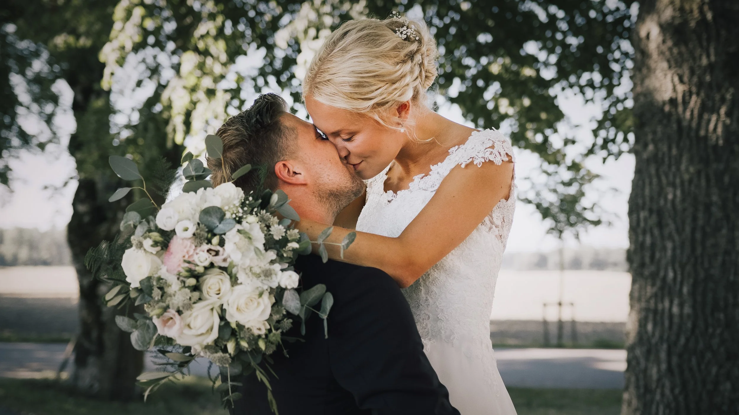 En brud och brudgummi pussas utomhus under ett träd. Bruden bär en vit brudklänning och håller en stor blomsterbukett, medan brudgummen har mörka kläder. Solsken filtreras genom bladen.