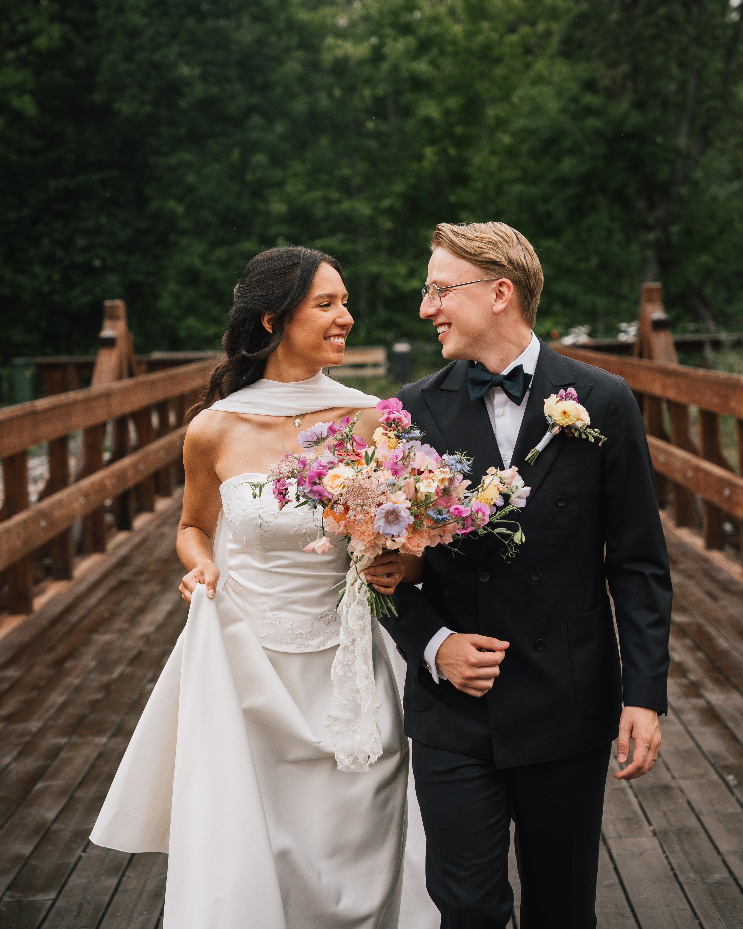 En nygift brud och brudgum som ler mot varandra på en träbro, medan bruden håller en stor bukett blommor. Bröllopskläder, civil bekvämlighet, gröna träd i bakgrunden.