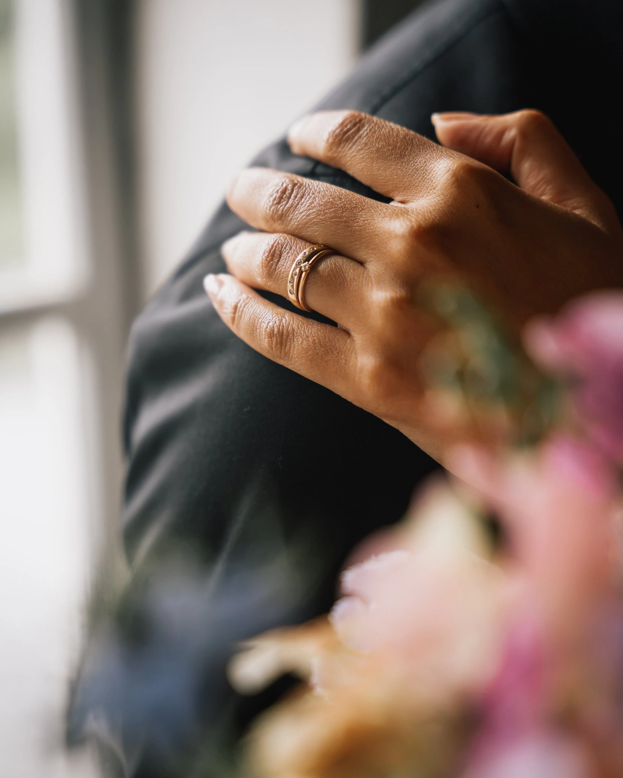 En hand med en guldring vilar på en svart yta, troligtvis en mans hand, med rosa blommor i förgrunden.