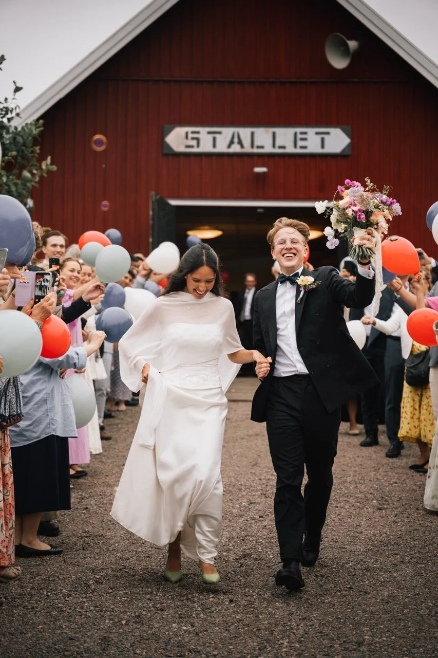 Nygift par som går hand i hand ut från ett rött stakspel, omringade av gäster med ballonger, utanför en lada med en skylt som säger "Stallet".