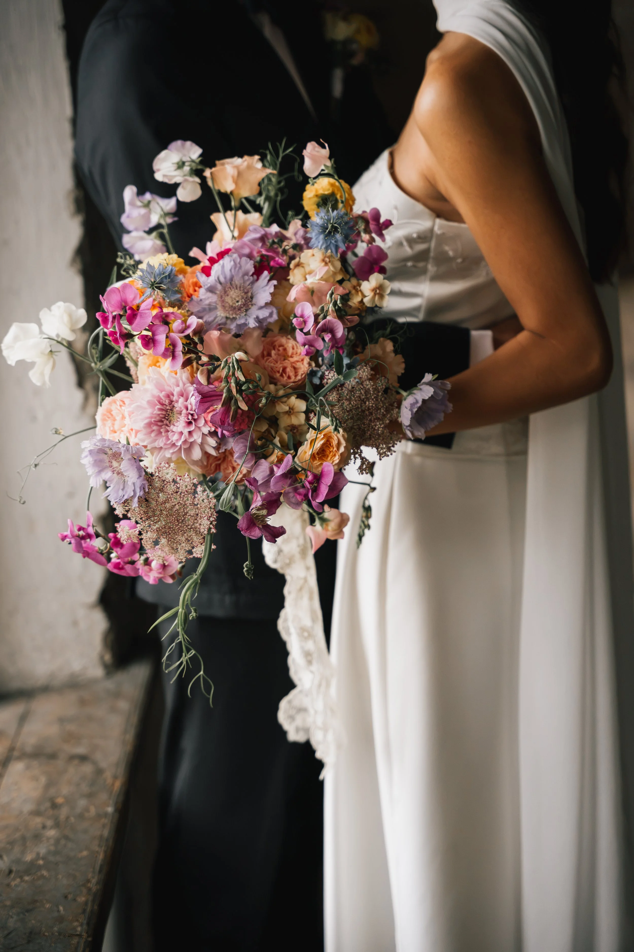 En person i brudklänning håller en stor bukett med blomster, hon står bredvid en person i svart kostym. Det är en bröllopsceremoni.