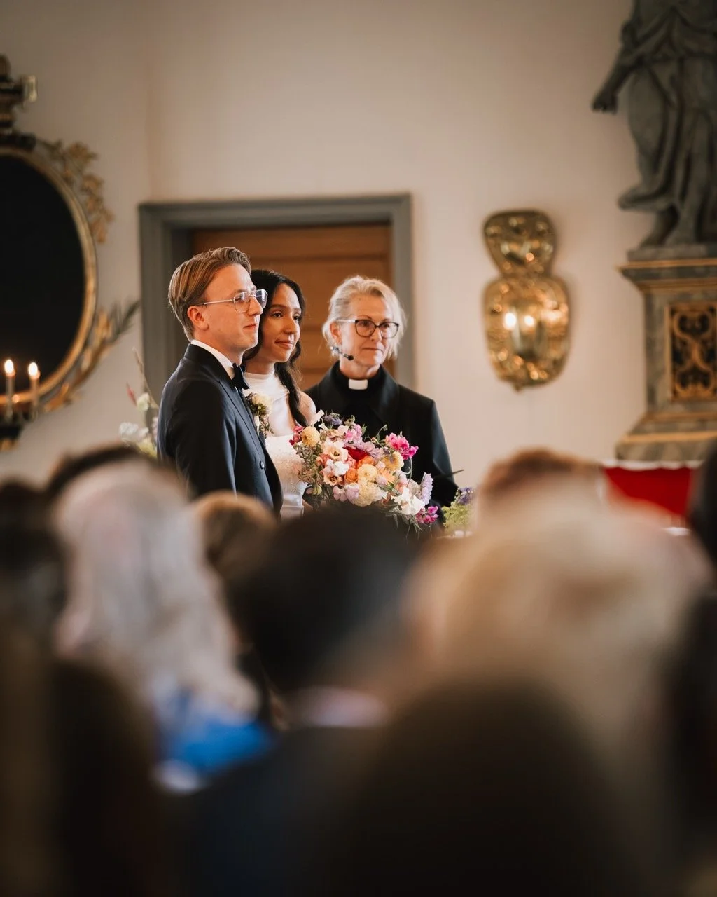Brudparet står tillsammans med en präst under ett bröllopsceremoni, omgiven av gäster i en kyrka.
