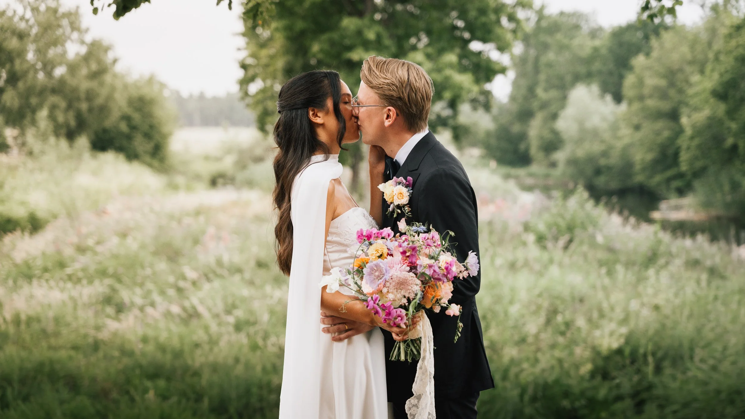 En brud och en brudgubb kysser varandra under ett utomhusbröllop, omgivna av gröna träd och ett blomsterhandlat bukett.