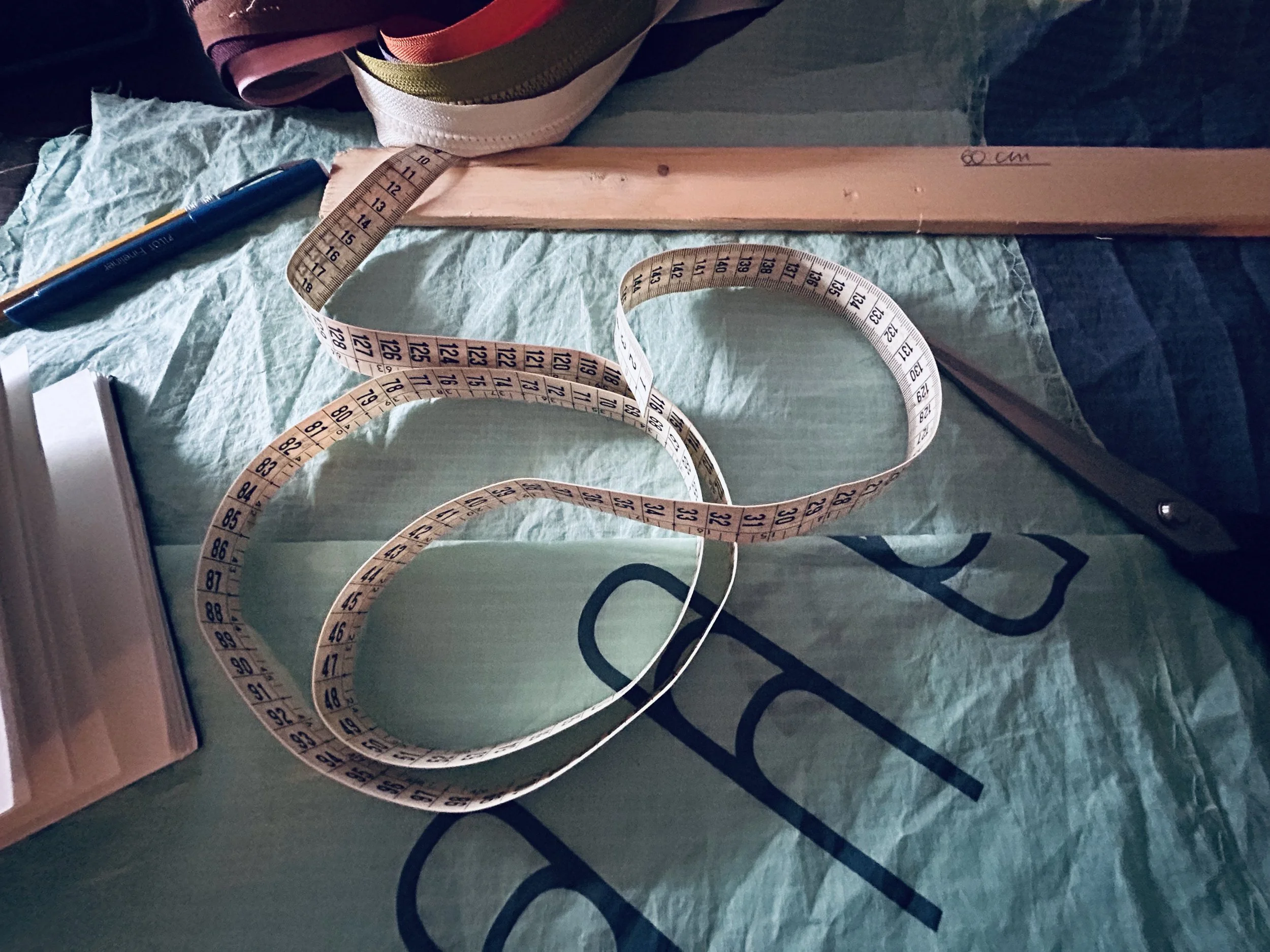 A blue marker, a measuring tape, and a piece of wood with markings are on a green surface, with fabric and a cardboard box nearby.