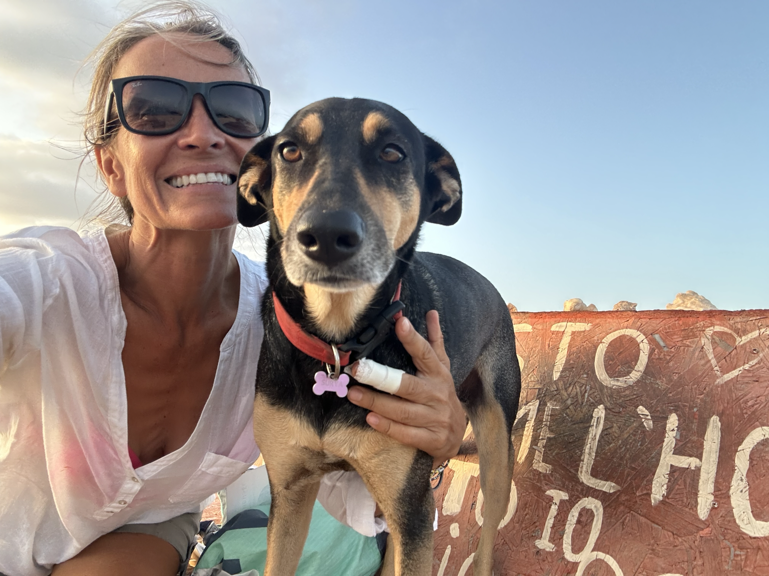 Smiling woman wearing sunglasses taking a selfie with her dog in front of a painted sign on a red bench during sunset.