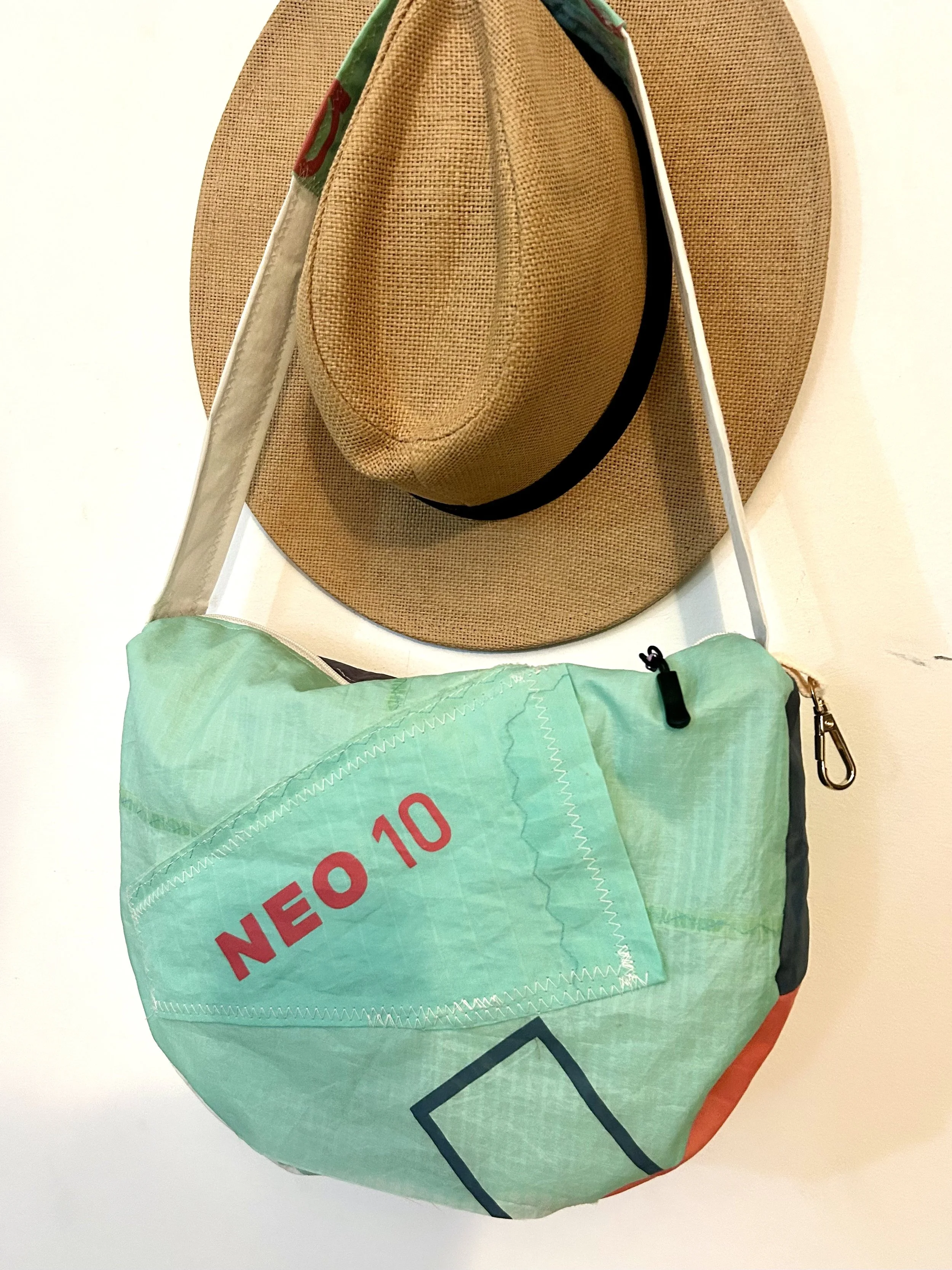 A aquamarine and purple half-moon Surya Le Sac bag hanging on a white wall, with a large brown tan hat with a black band hanging behind it.