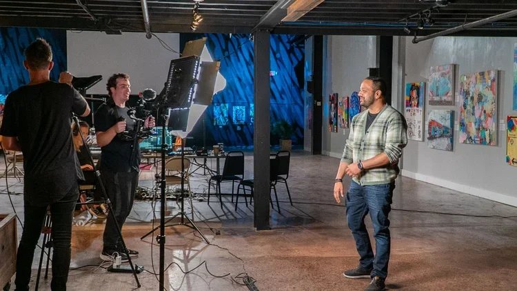 Professional lighting and camera on the left pointing towards and man at the right. There are two Helm team members working behind the setup.