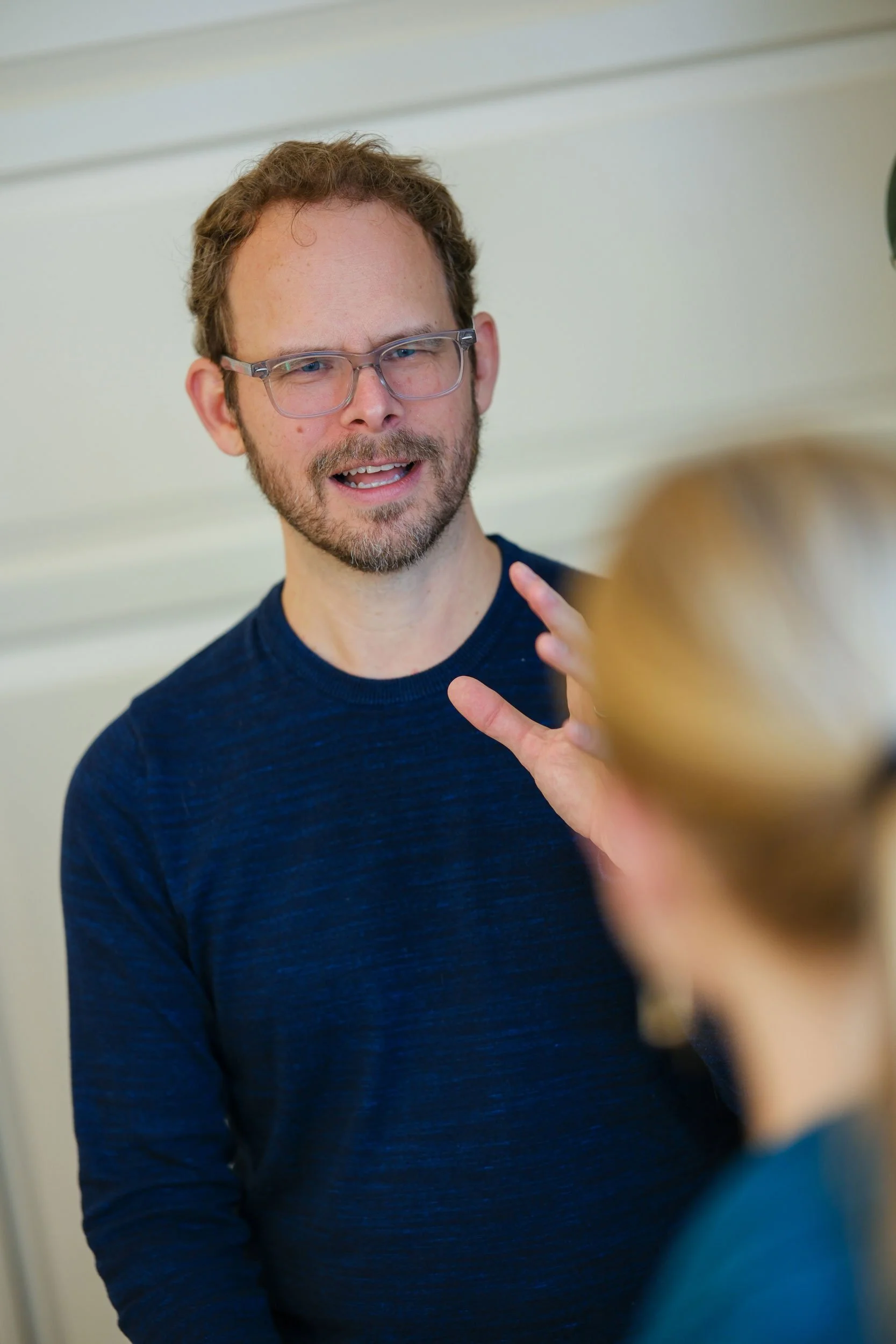 Adam Sippola, a man with glasses and a beard, wearing a dark blue shirt, is talking and gesturing with his hand while conversing with a woman whose face is out of focus.