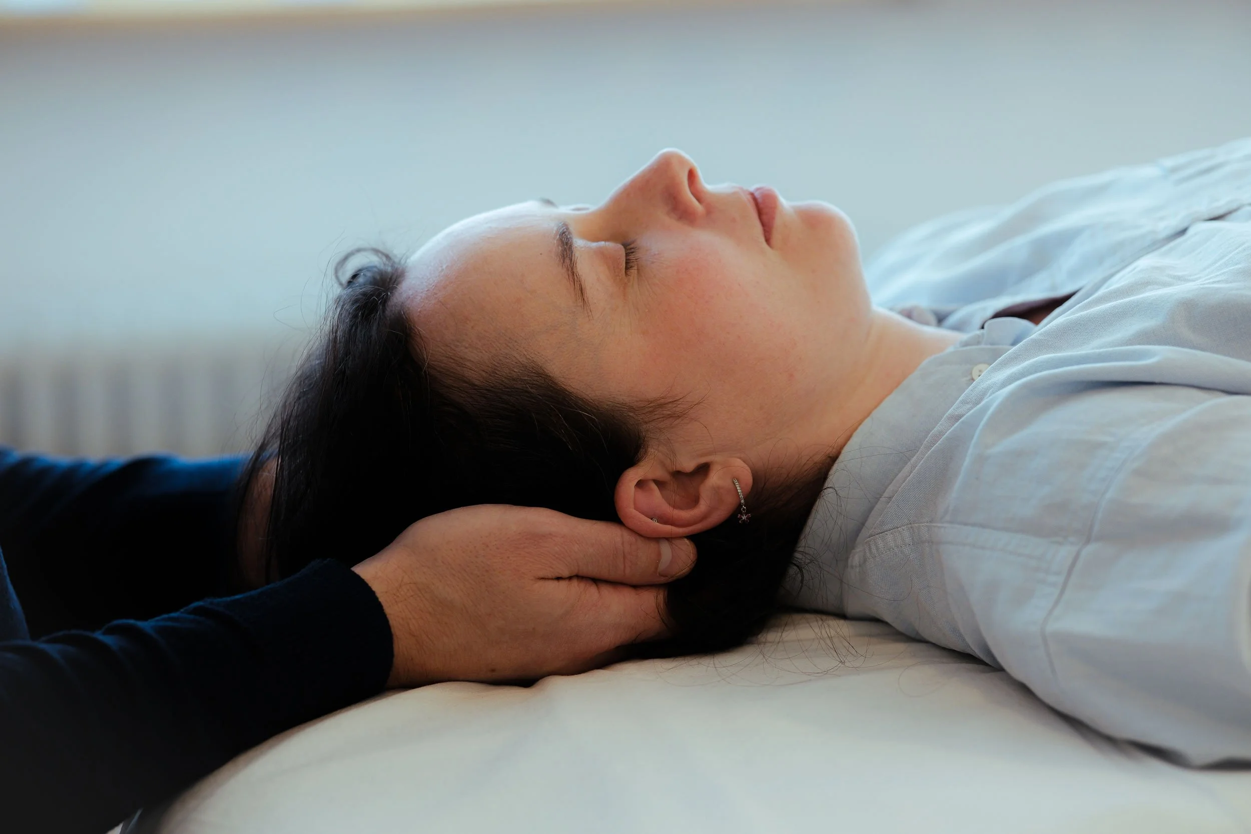 A woman lying on her back with eyes closed, receiving a Craniosacral Therapy session. Hands hold her neck and head as she lays on a massage table.