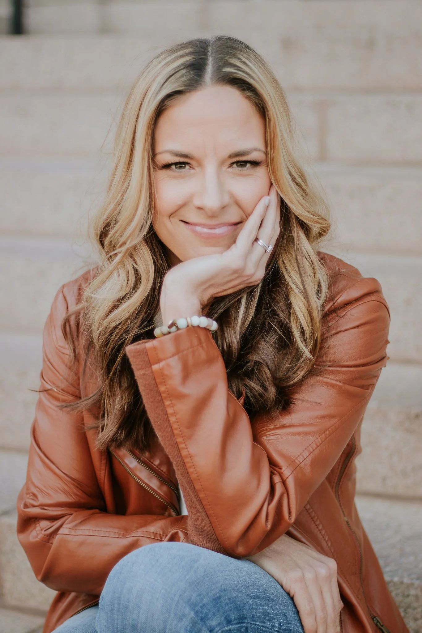 A woman with long, wavy blonde hair smiling, resting her face on her hand, wearing a brown leather jacket and blue jeans, seated outdoors in front of beige brick steps.