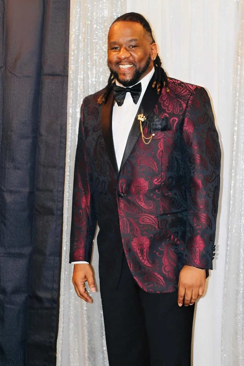 A man wearing a dark red and black paisley patterned tuxedo jacket, white shirt, black bow tie, and black pants, standing in front of a silver and black curtain, smiling at the camera.