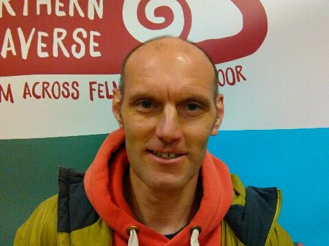 A man wearing a red hoodie and dark jacket, standing in front of a colorful wall with red, blue, and green sections and writing on it.