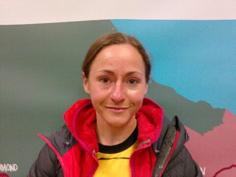 A woman with brown hair wearing a black and red jacket smiling in front of a colorful map.