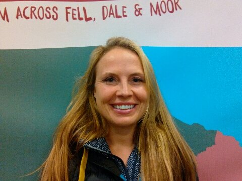 A smiling woman with long blonde hair standing in front of a colorful wall with text and graphics.