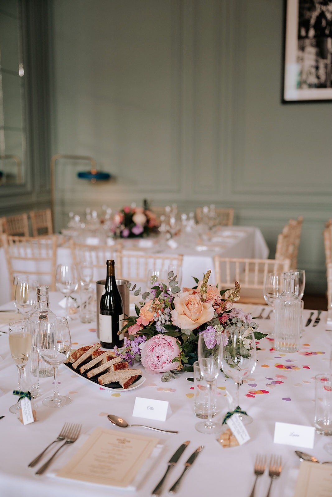 A decorated banquet table with a floral centerpiece, wine, glasses, plates, cutlery, a menu, and small name tags in a decorated room.