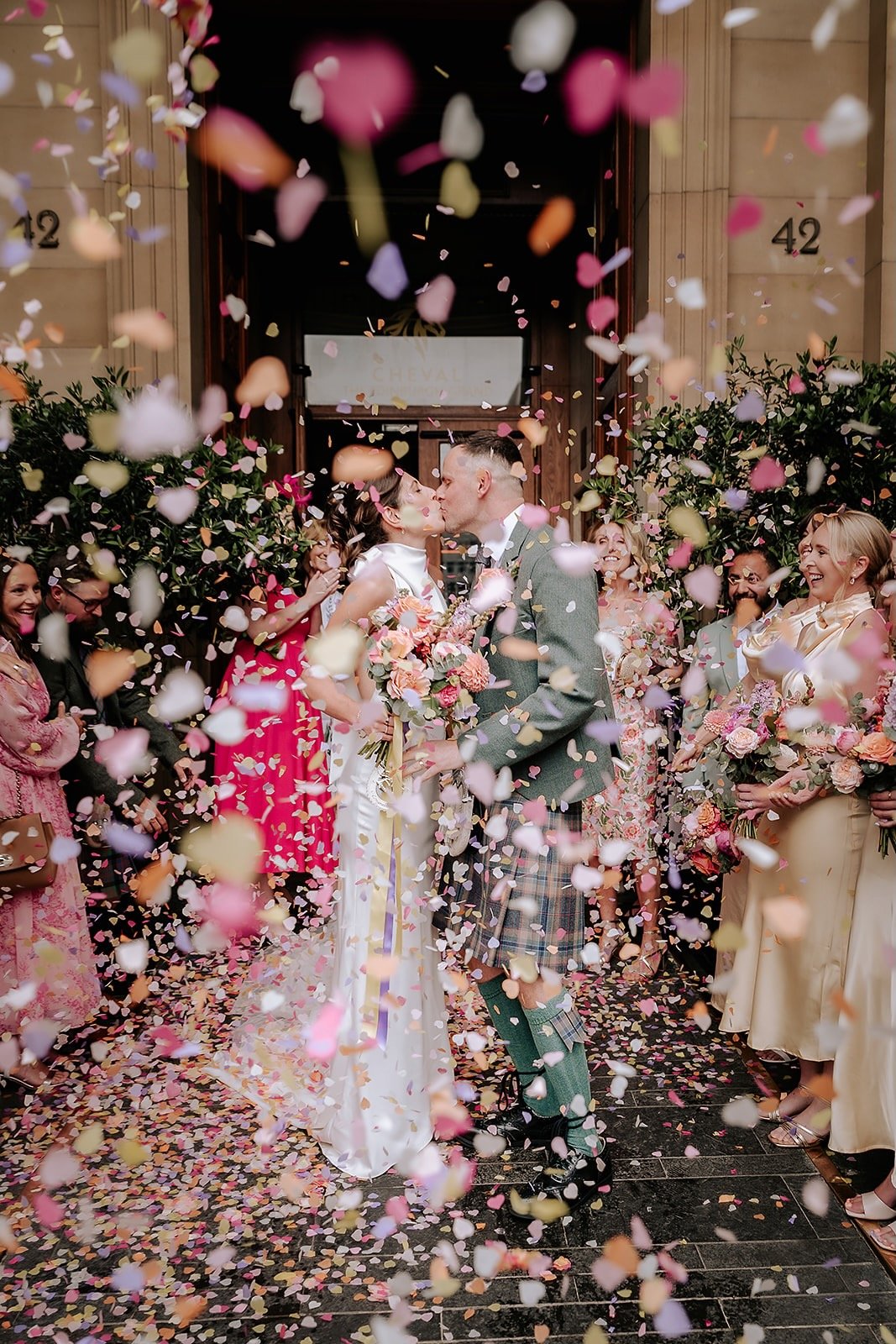 A bride and groom kiss as they are showered with colorful confetti, surrounded by smiling friends and family outside a building decorated for a wedding.