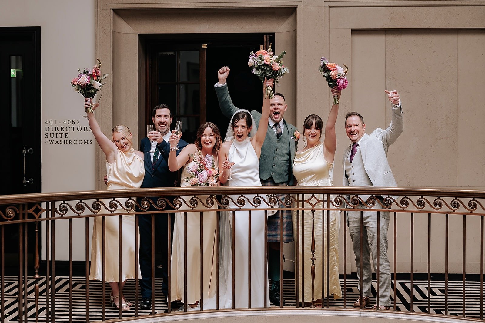 A group of wedding celebration guests celebrating on a balcony with bouquets and champagne, surrounded by a beige indoor setting.